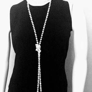 May include: A long, white beaded necklace with a knotted centerpiece hangs over a black sleeveless dress. The necklace is composed of small, round beads and creates a Y-shape. The image is in black and white.
