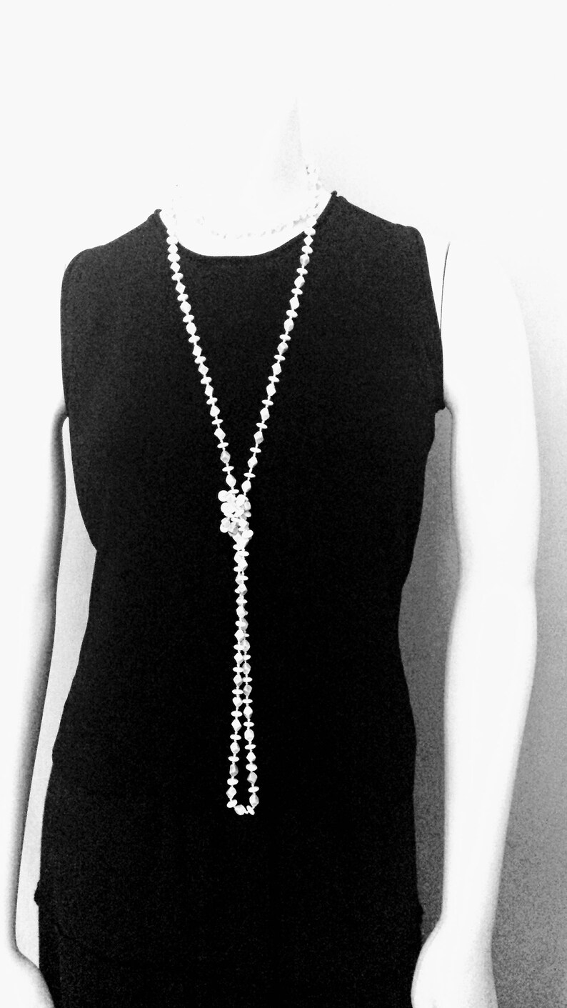 May include: A long, white beaded necklace with a knotted centerpiece hangs over a black sleeveless dress. The necklace is composed of small, round beads and creates a Y-shape. The image is in black and white.