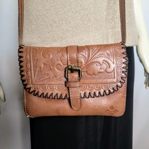 May include: A brown leather crossbody bag with a floral embossed design. The bag features a buckle closure and a black stitched border. The strap is brown, and the bag has a small logo on the bottom right corner.