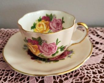 Puede incluir: Un juego de taza y platillo de té vintage. La taza y el platillo son de color rosa pálido con un ribete dorado. La taza está decorada con un estampado floral de rosas amarillas, rosas y rojas. El platillo tiene un diseño floral a juego.