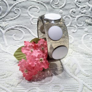 May include: A clear, glitter-infused acrylic bangle bracelet with three large, white, circular accents. The bracelet is displayed with a sprig of pink flowers and green leaves, set against a patterned white background.
