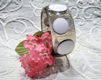 May include: A clear, glitter-infused acrylic bangle bracelet with three large, white, circular accents. The bracelet is displayed with a sprig of pink flowers and green leaves, set against a patterned white background.