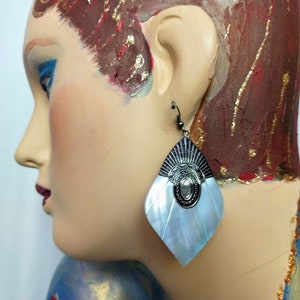 May include: A close-up of a mannequin wearing a pair of earrings. The earrings have a silver-colored top with a central oval design and a pearlescent, fan-shaped bottom. The mannequin has red lipstick and blue eyeshadow.