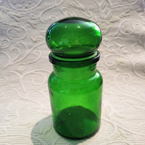 Puede incluir: Un tarro de boticario de vidrio verde brillante con tapa redondeada. El tarro tiene un cuerpo cilíndrico y un borde negro. La tapa es verde a juego, creando una pieza cohesiva y decorativa. El tarro probablemente se usa para almacenamiento.