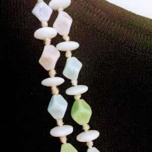 May include: A pastel-colored necklace featuring alternating shapes of white, light blue, pink, and green beads. The beads are strung together with a beige cord, creating a delicate and vintage aesthetic. The necklace is displayed against a dark background.