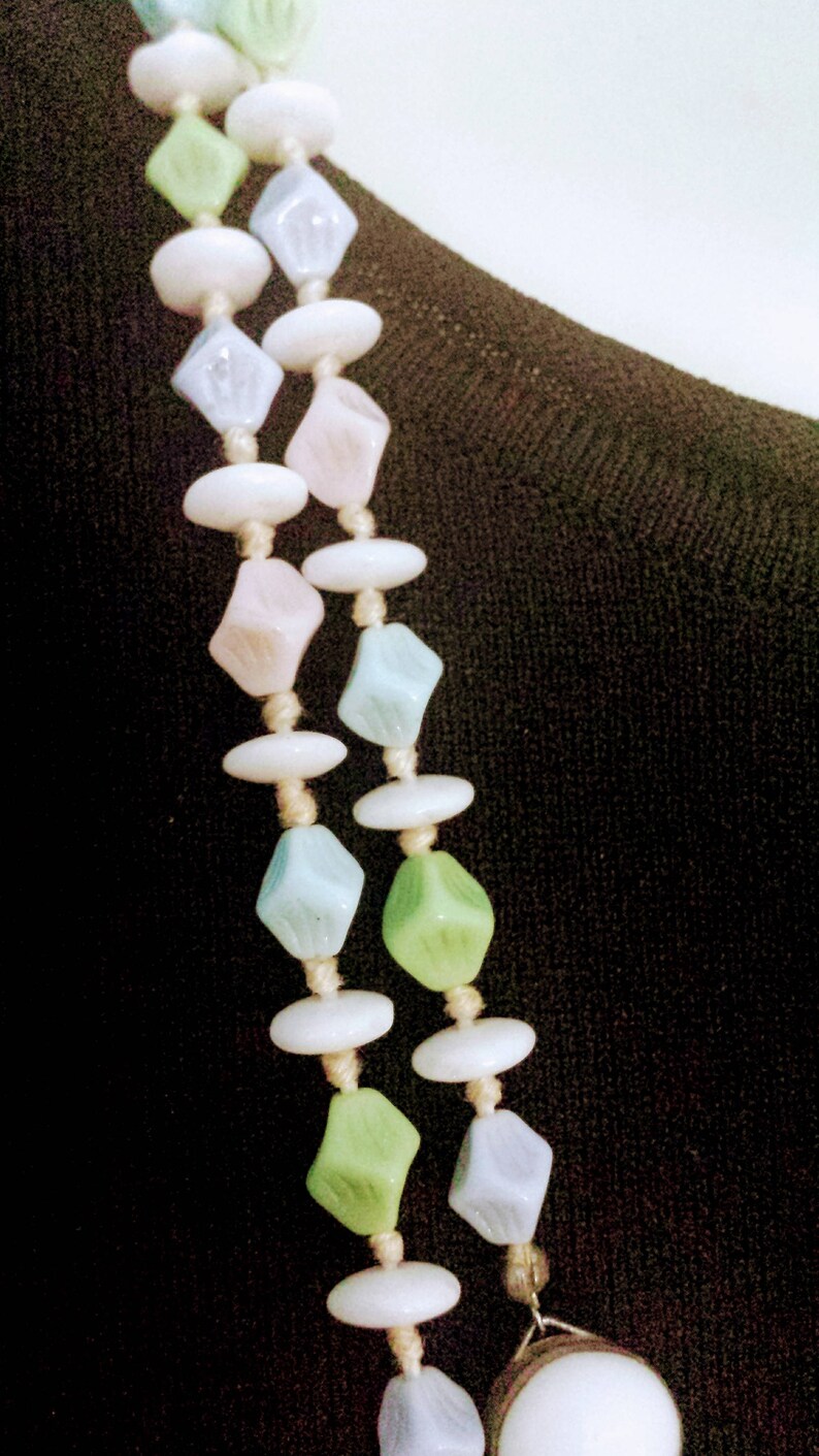 May include: A pastel-colored necklace featuring alternating shapes of white, light blue, pink, and green beads. The beads are strung together with a beige cord, creating a delicate and vintage aesthetic. The necklace is displayed against a dark background.