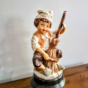 May include: A ceramic figurine of a young boy in a white chef's hat and brown vest, holding a broom. The figurine has a detailed face and is mounted on a black base. The boy is looking upwards.