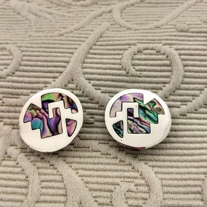 May include: Pair of silver-toned stud earrings. Each earring features a circular design with a cut-out shape filled with iridescent abalone shell. The earrings are displayed on a textured, light gray surface.