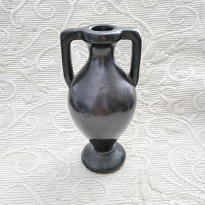 May include: A dark gray ceramic vase with two handles and a pedestal base. The vase has a smooth, glossy finish and a small opening at the top. The vase is approximately 6 inches tall.