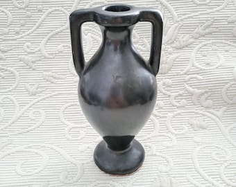 Può includere: Un vaso in ceramica grigio scuro con due manici e una base a piedistallo. Il vaso ha una finitura liscia e lucida e una piccola apertura nella parte superiore. Il vaso è alto circa 15 cm.