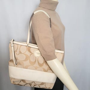 May include: Beige and cream Coach tote bag with a repeating logo pattern. The bag features cream-colored handles and a cream-colored stripe across the bottom. A circular Coach logo is visible on the front.