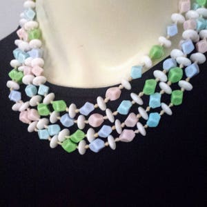 May include: A multi-strand necklace featuring pastel-colored beads. The necklace has three strands of cube-shaped beads in shades of pink, blue, and green, interspersed with white oval beads. The necklace is displayed on a mannequin.