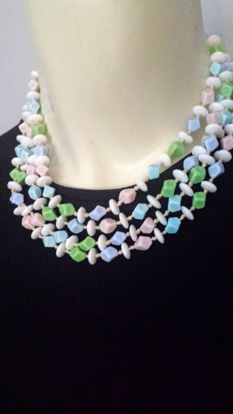 May include: A multi-strand necklace featuring pastel-colored beads. The necklace has three strands of cube-shaped beads in shades of pink, blue, and green, interspersed with white oval beads. The necklace is displayed on a mannequin.