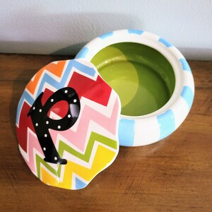 May include: A ceramic trinket box with a lid featuring a colorful zigzag pattern in red, orange, pink, green, and yellow. The lid has a black letter "P" with white polka dots. The box is white with blue stripes.