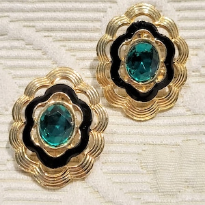 May include: A pair of gold-toned earrings with a floral design. Each earring features a black enamel border and a central, oval-shaped, emerald-green gemstone. The earrings are set against a textured, off-white background.