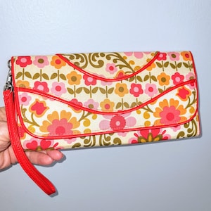 May include: A floral print clutch with a red wrist strap. The clutch features a cream-colored background with a repeating pattern of pink, orange, and yellow flowers. Red trim outlines the flap and edges of the clutch.