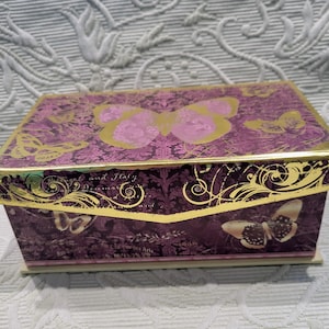 Puede incluir: Una caja decorativa con un diseño de mariposas moradas y doradas. La caja presenta un patrón de mariposas repetido y adornos dorados. La tapa tiene una mariposa grande en el centro. La caja es rectangular y tiene una base de color claro.