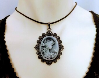 May include: Oval cameo pendant necklace featuring a profile of a woman with flowing hair and floral accents. The pendant is set in a decorative, dark metal frame and hangs from a dark brown cord necklace. The cameo is a light gray color.