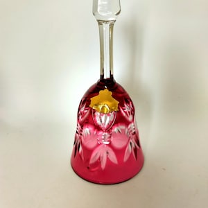May include: A ruby red crystal bell with a clear, faceted handle and clapper. The bell features a decorative cut pattern and a gold-colored sticker. The bell is a decorative item.