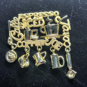 Pode incluir: Broche dourado com tema de café com as palavras "Espresso", "Iced", "Coffee" e "Joe". O broche apresenta mini xícaras de café, uma colher e um pingente de cafeteira. O broche está em um fundo preto.