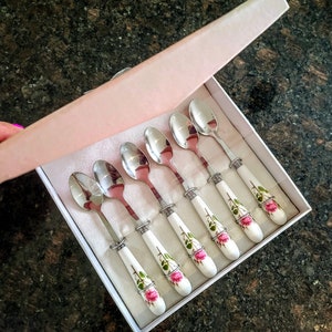 Könnte beinhalten: Ein Set aus sechs silberfarbenen Teelöffeln mit weißen Porzellangriffen, die mit rosa Rosen und grünen Blättern verziert sind, präsentiert in einer weißen Schachtel mit einem rosa Deckel.