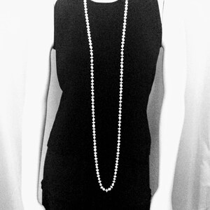 May include: A sleeveless black dress with a long, white beaded necklace. The necklace hangs down the front of the dress. The image is in black and white.