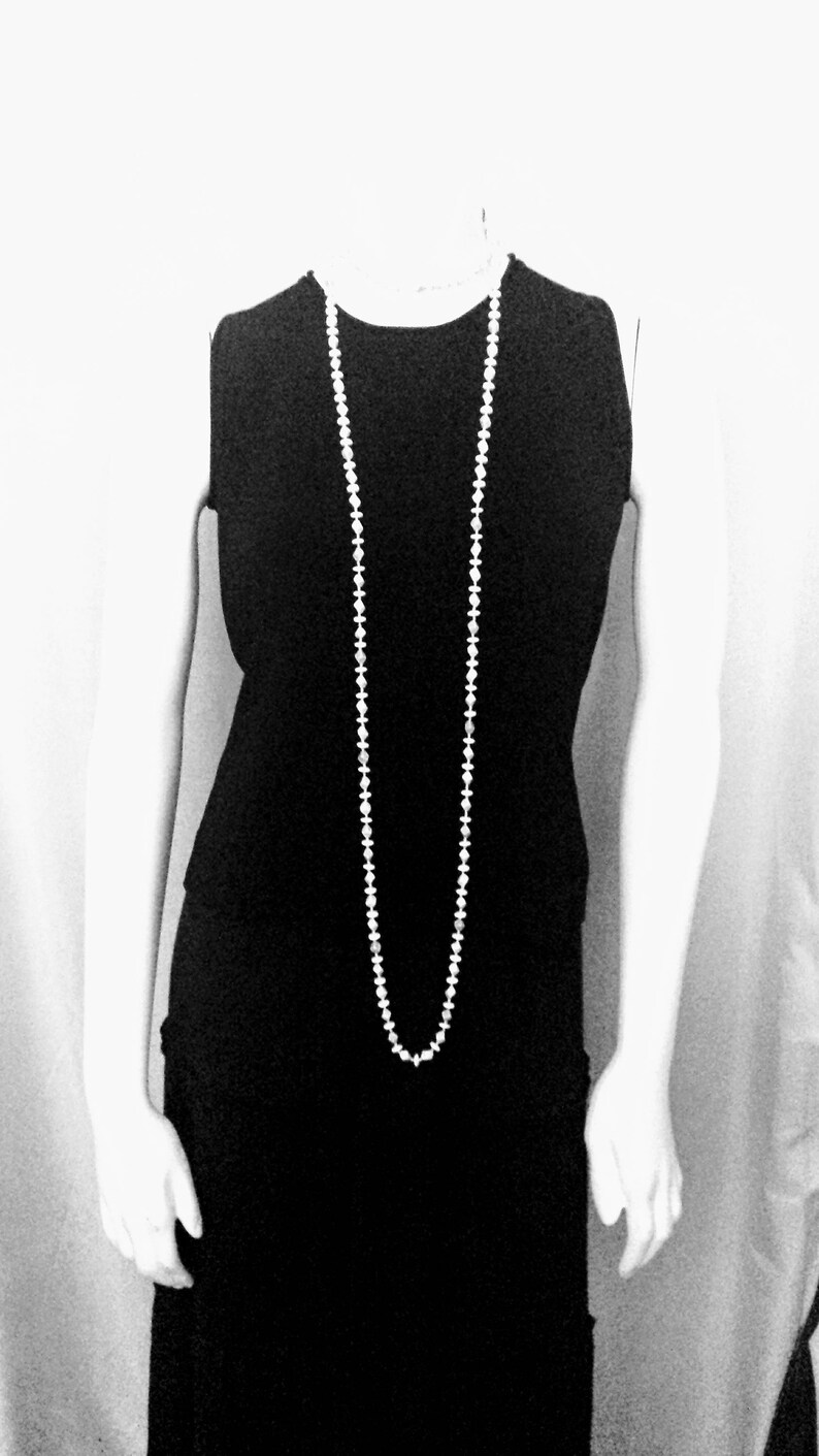 May include: A sleeveless black dress with a long, white beaded necklace. The necklace hangs down the front of the dress. The image is in black and white.