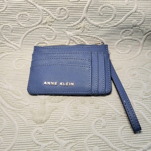 Puede incluir: Un tarjetero Anne Klein azul bígaro con una correa de muñeca. El tarjetero tiene múltiples ranuras para tarjetas y un compartimento con cremallera. El logotipo de Anne Klein está en letras doradas.