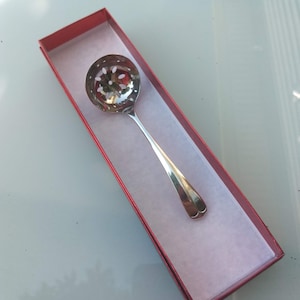 May include: A silver slotted spoon with a round bowl and long handle, presented in a red rectangular gift box. The spoon's bowl features a floral pattern of holes. The box is lined with white paper.