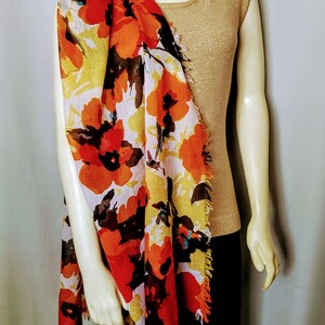 May include: A floral scarf with red, orange, yellow, and brown flowers is draped over a gold-colored top. The scarf has a white background and fringed edges. The mannequin is wearing a black skirt.