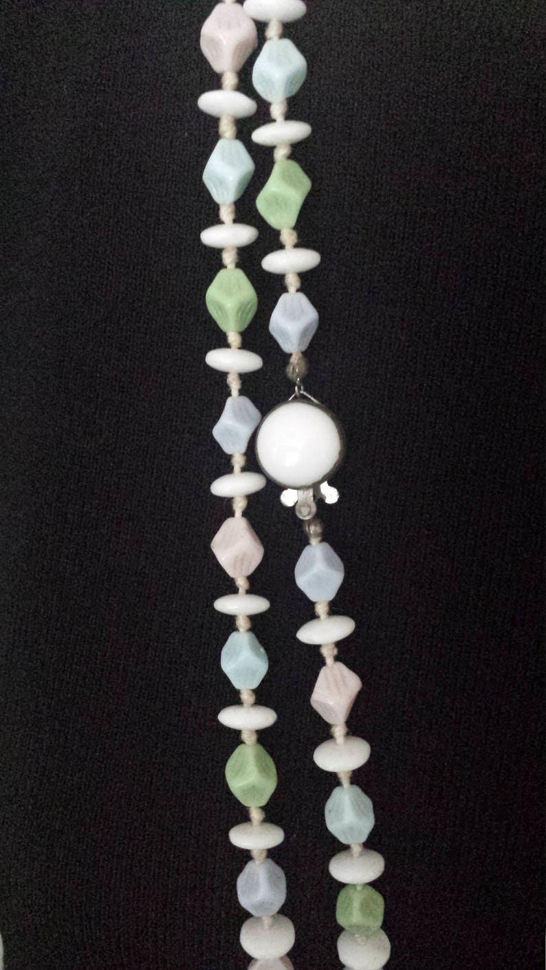 May include: A vintage necklace featuring two strands of beads in pastel colors. The beads include diamond-shaped and oval beads in shades of pink, blue, green, and white. A large white circular pendant is attached.