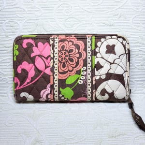 Puede incluir: Un monedero acolchado marrón con un estampado floral en rosa, verde y blanco. El monedero tiene cierre de cremallera y una correa de muñeca. La marca Vera Bradley es visible en el lateral.