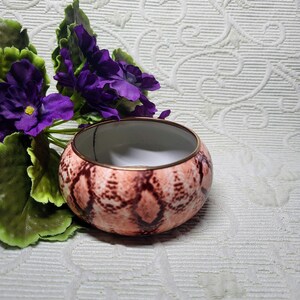 May include: A small, round decorative bowl with a snakeskin pattern in shades of pink and brown. The rim is trimmed with a gold-colored metal. The bowl is next to a cluster of purple flowers and green leaves.