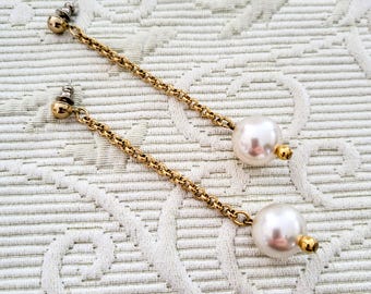 May include: A pair of dangle earrings featuring a gold-colored chain and a large, round, white pearl. The earrings have a post-back closure and a small gold-colored bead at the base of the pearl. The earrings are displayed on a white, textured surface.