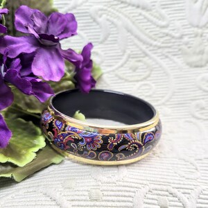 May include: A black bangle bracelet with a gold rim and a colorful paisley design in red, blue, and purple. The bracelet is set against a backdrop of purple flowers and a white patterned surface.
