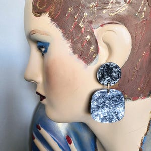 May include: Statement earrings with a marbled design. Each earring has a small, circular top and a larger, square bottom, joined by a gold-tone link. The marbled effect combines black and white, creating a unique accessory.