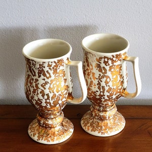 Puede incluir: Dos tazas vintage con pedestal, base color crema y un patrón moteado dorado. Cada taza tiene un asa y un borde ancho. Las tazas se exhiben sobre una superficie de madera contra una pared gris claro.