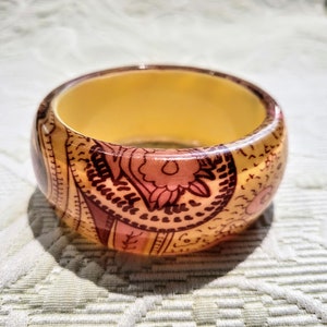 May include: A wide, round bangle bracelet with a warm, amber-colored interior. The exterior is decorated with a repeating pattern of floral and paisley designs in shades of brown and pink. The bracelet is made of a glossy material.