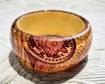May include: A wide, round bangle bracelet with a warm, amber-colored interior. The exterior is decorated with a repeating pattern of floral and paisley designs in shades of brown and pink. The bracelet is made of a glossy material.