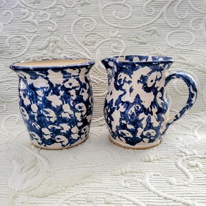 May include: Two ceramic pieces with a blue and white floral pattern. One is a small pitcher with a handle, and the other is a cylindrical vase. Both have a rustic, handmade appearance and are set against a white background.