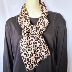 May include: A tan and brown leopard print scarf is draped around a mannequin's neck. The scarf has a fringed edge and is knotted at the front. The mannequin is wearing a long-sleeved black shirt.