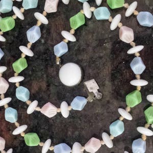 May include: A pastel-colored necklace with a variety of beads. The necklace features light blue, pink, and green cube-shaped beads, interspersed with white oval beads. The necklace is secured with a silver clasp.