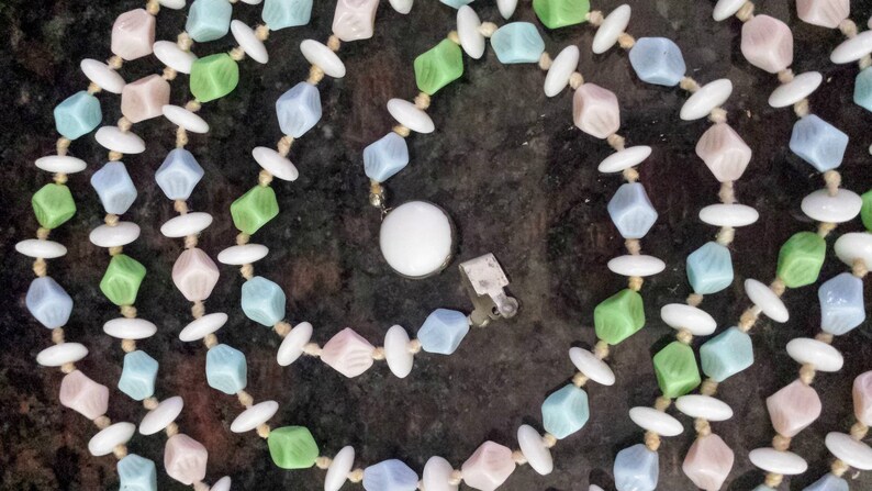 May include: A pastel-colored necklace with a variety of beads. The necklace features light blue, pink, and green cube-shaped beads, interspersed with white oval beads. The necklace is secured with a silver clasp.