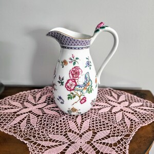 May include: A white ceramic pitcher with a floral and butterfly design. The pitcher has a gold rim and a handle with a pink rose accent. The base of the pitcher is resting on a pink lace doily.