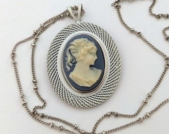 May include: A silver-toned necklace featuring an oval pendant with a cameo profile. The cameo is set against a blue background, framed by a white border and a mesh-like silver surround. The chain is delicate and beaded.