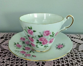 May include: White teacup and saucer set with pink rose floral pattern and gold trim. The teacup has a curved handle and sits on a matching saucer. The set is likely made of porcelain and is suitable for serving tea.