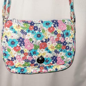 May include: A floral crossbody bag with a quilted pattern. The bag displays a vibrant mix of colourful flowers in shades of blue, pink, yellow, and red against a white background. It has a silver zip and a black button closure.