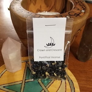 Puede incluir: Una pequeña bolsa de plástico transparente que contiene incienso negro y dorado. La bolsa tiene una etiqueta blanca con el texto "Crown and Crescent" y "Pontifical Incense".