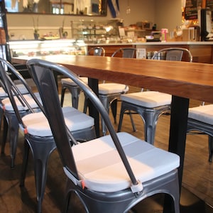 May include: A long, rectangular wooden table with a dark finish, supported by black metal legs. Surrounding the table are several metal chairs with gray seat cushions. The chairs have a classic industrial design, suitable for a cafe or restaurant setting.
