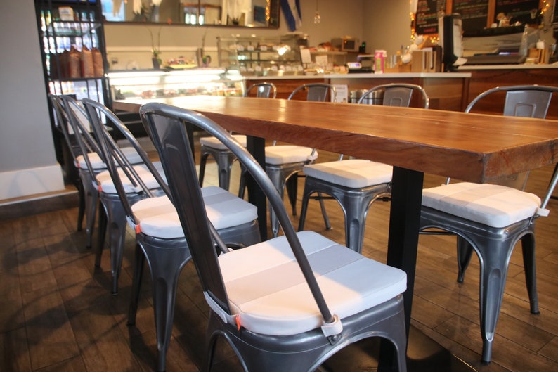 May include: A long, rectangular wooden table with a dark finish, supported by black metal legs. Surrounding the table are several metal chairs with gray seat cushions. The chairs have a classic industrial design, suitable for a cafe or restaurant setting.